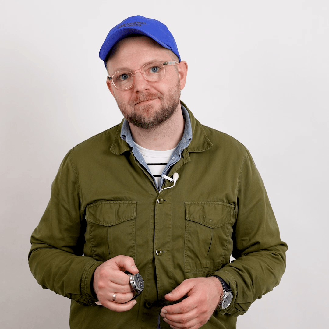 Bearded man in glasses and blue cap holding a wristwatch, dressed in an olive overshirt over a striped shirt against a neutral background