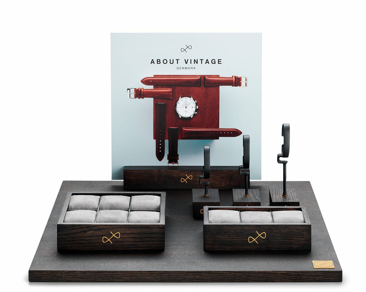 About Vintage watch display stand with dark wood trays, gray cushions, upright leather straps, and a coordinating wall graphic