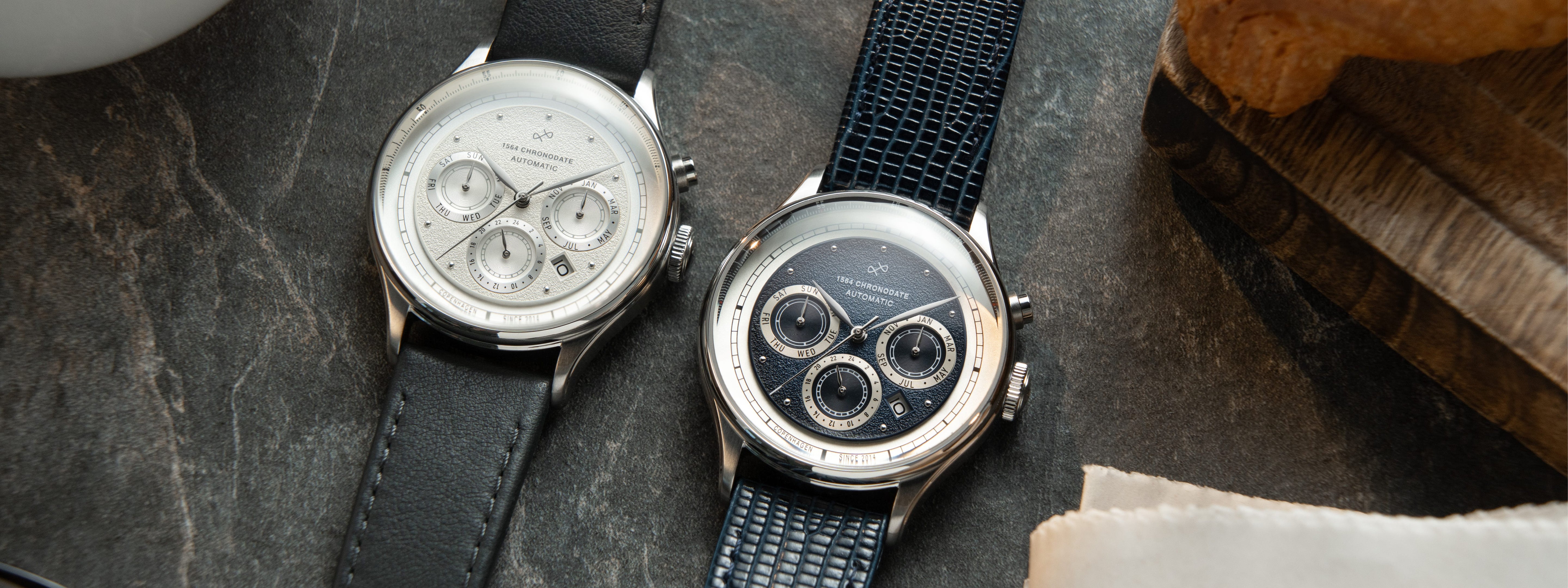 Two AV86 chronograph watches with textured dials, subdials for day, month, and 24-hour time, sapphire crystals, and leather straps, displayed on a dark stone surface alongside wooden and fabric elements, reflecting Nordic minimalism and Danish horological precision.