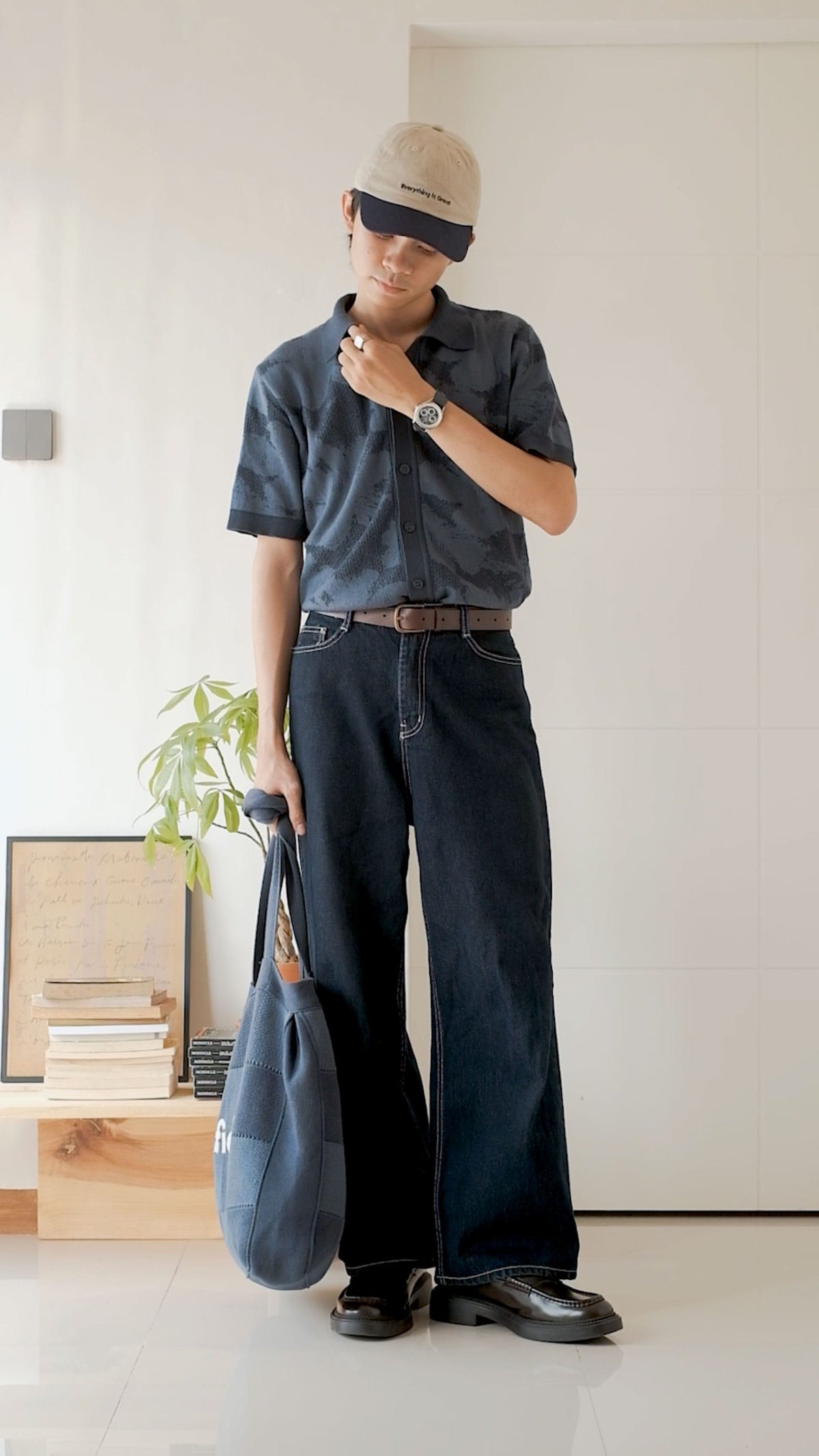 Person in a relaxed indoor setting wearing a cap, dark patterned shirt, wide-leg trousers, and a minimalist wristwatch with a round dial.