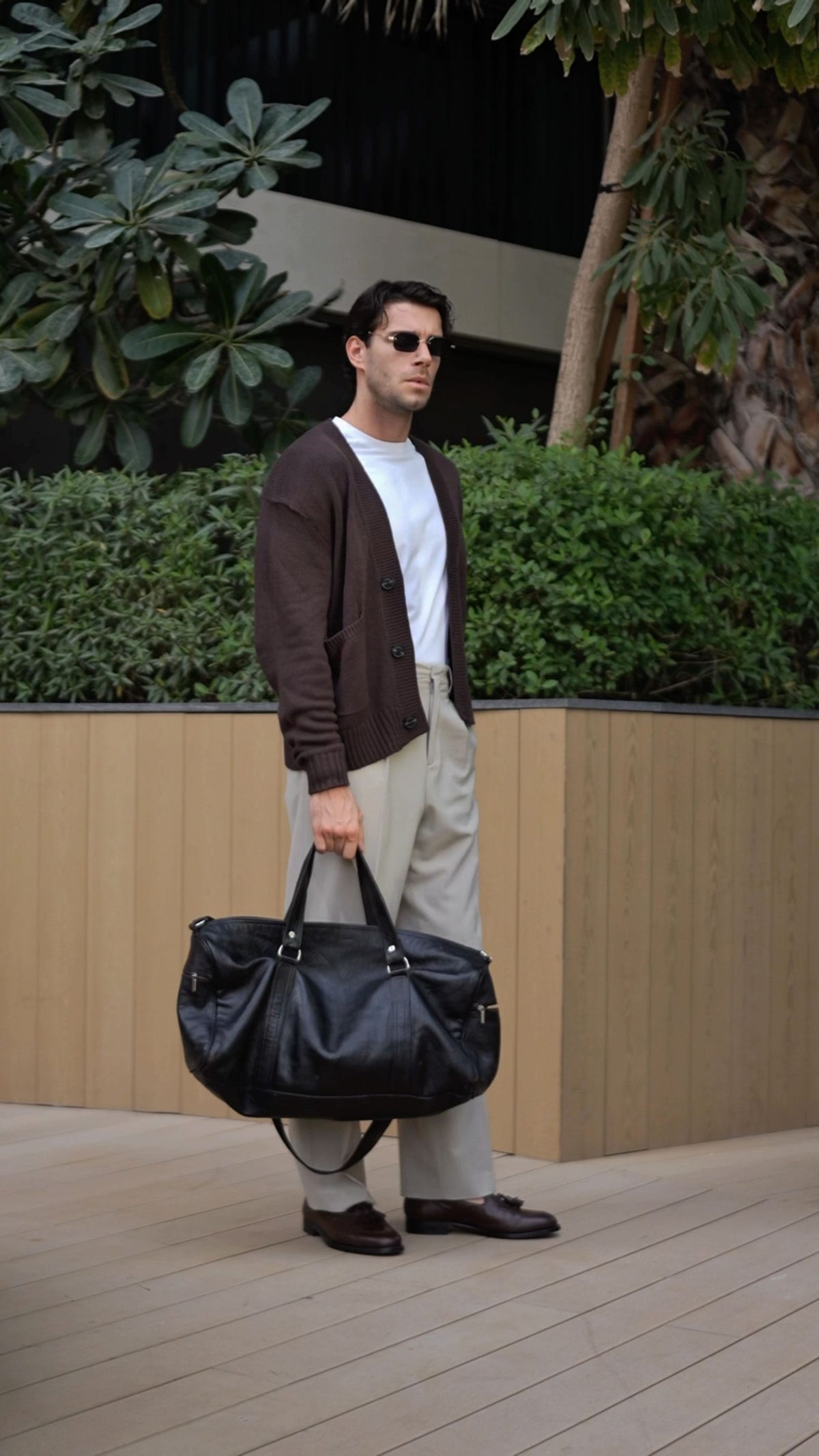 Man in sunglasses and a dark cardigan carrying a black leather weekender bag beside greenery in a modern outdoor setting