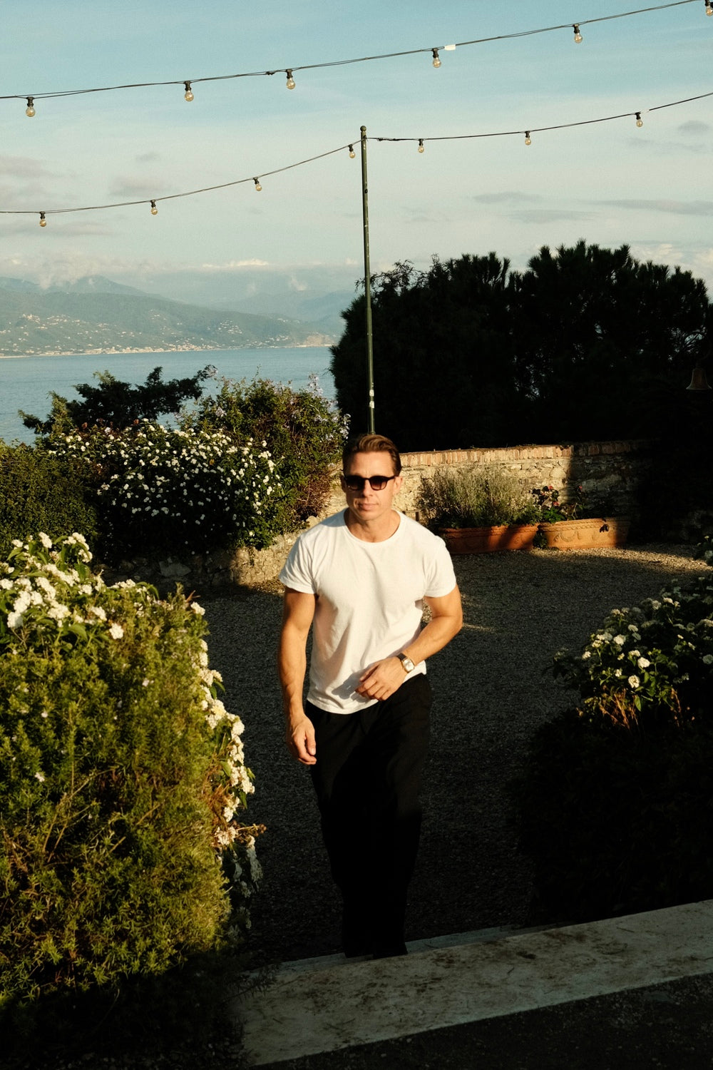 Man wearing sunglasses and a 1954 Amalfi Mechanical, Steel / Off White watch by AV86 walking outdoors near greenery and string lights.