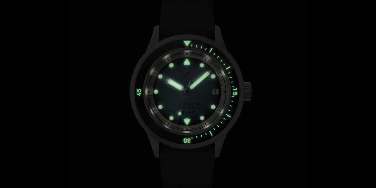 Diver style wristwatch glowing green luminescent hands and markers on a dark dial with a black strap.