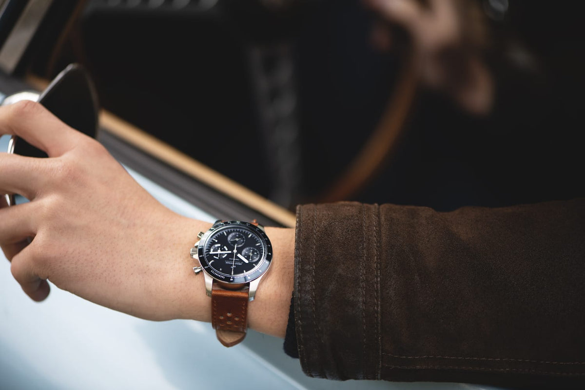1960 Racing Chronograph by AV86 with vintage-inspired dial, sapphire crystal, and brown leather strap on wrist driving a car
