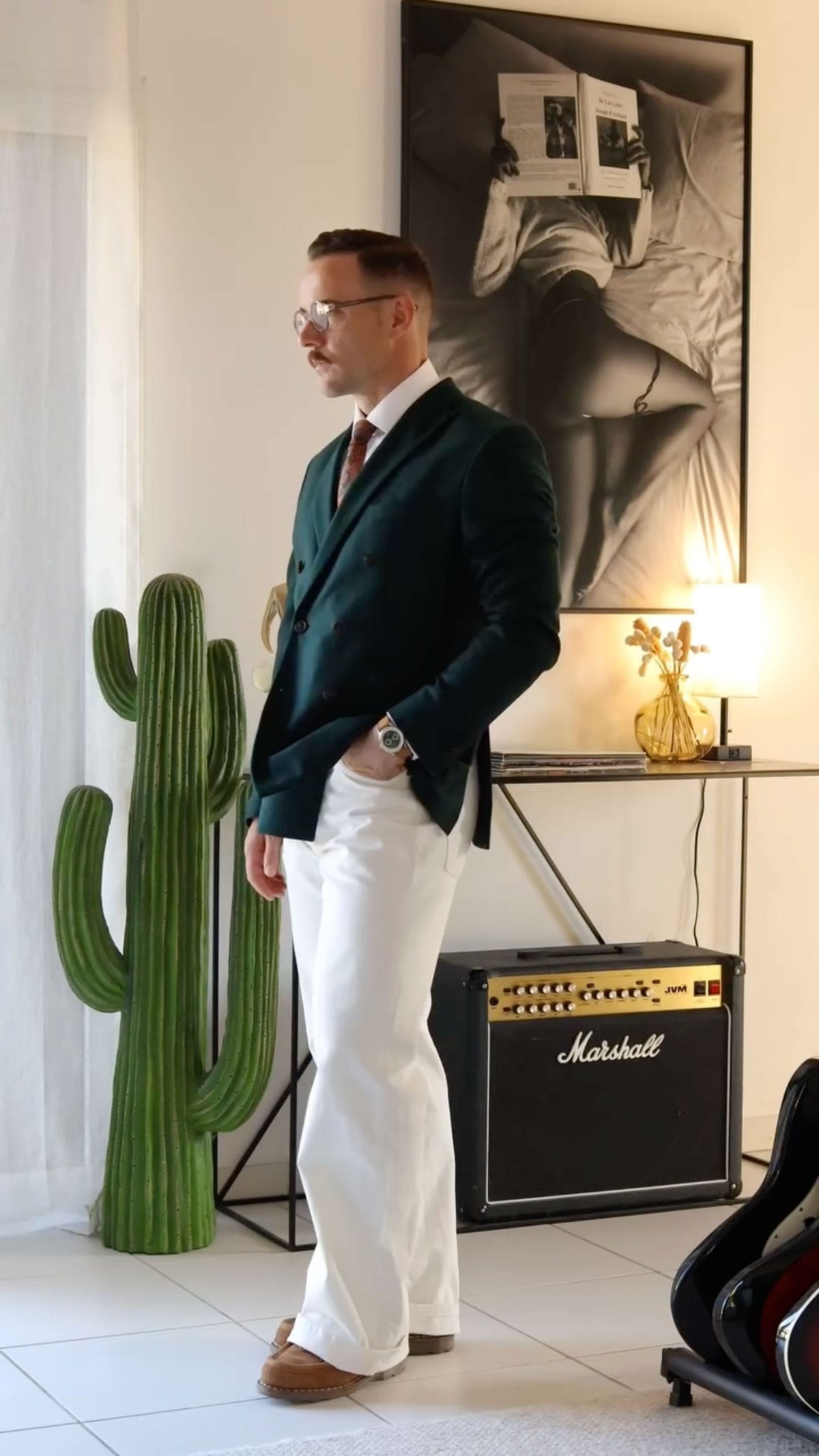 Man in a dark tailored suit and white trousers wearing a wristwatch, standing beside a cactus and Marshall amplifier in a refined interior