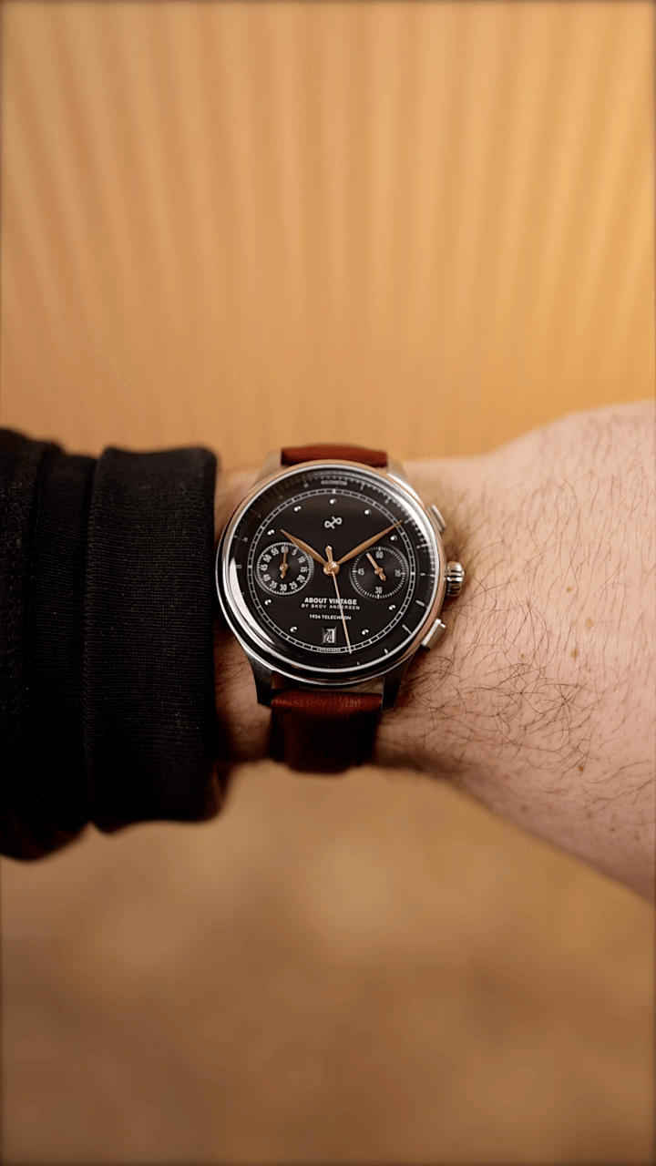 AV86 vintage-inspired chronograph with dark dial, twin subdials, polished case and brown leather strap worn against a warm wood backdrop