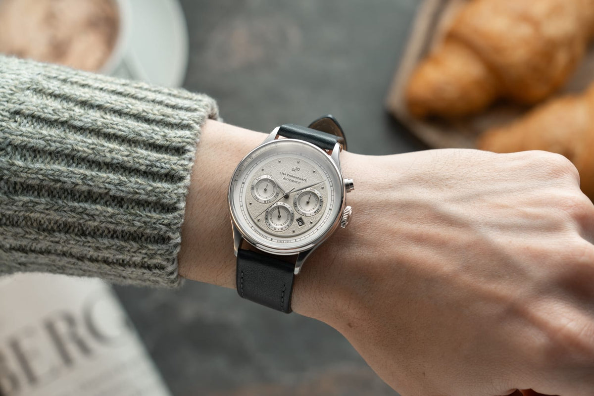 1564 Chronodate Automatic by AV86 displays a vintage-inspired automatic chronograph with a textured dial, triple subdials, polished stainless steel case, sapphire crystal, and black leather strap, worn in a cozy setting featuring soft knitwear and baked goods, embodying Nordic minimalism and Danish horological excellence.