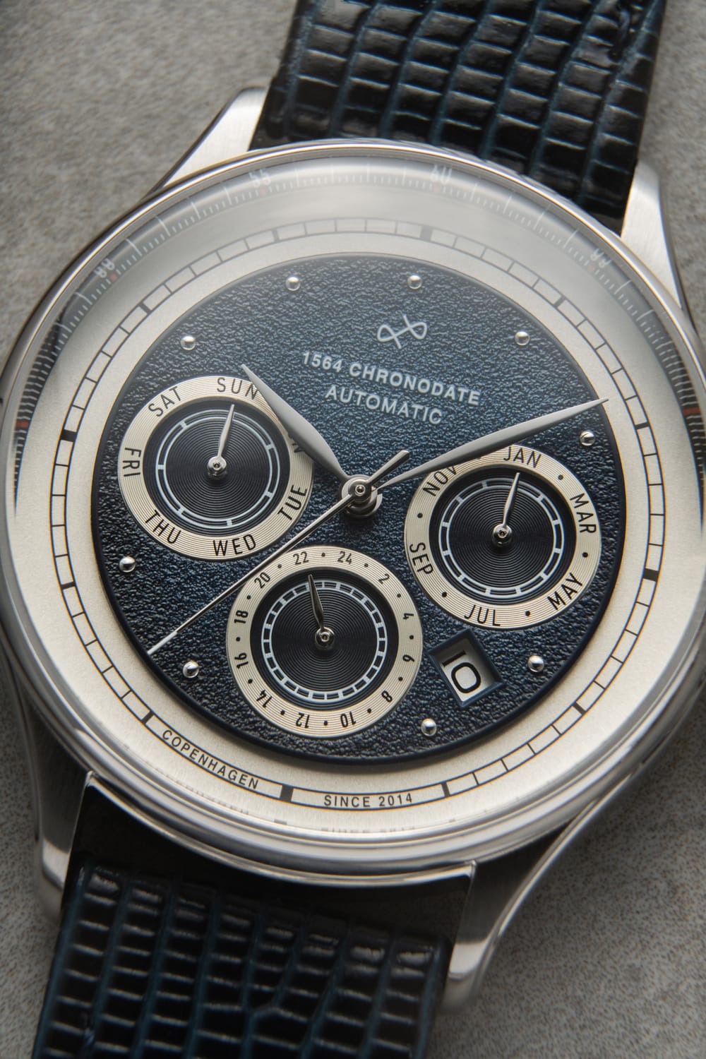 1564 Chronodate Automatic by AV86 features a textured dial with day, month, and 24-hour subdials, date window, polished case, and a black patterned leather strap, exemplifying Nordic minimalism and Danish horological precision.