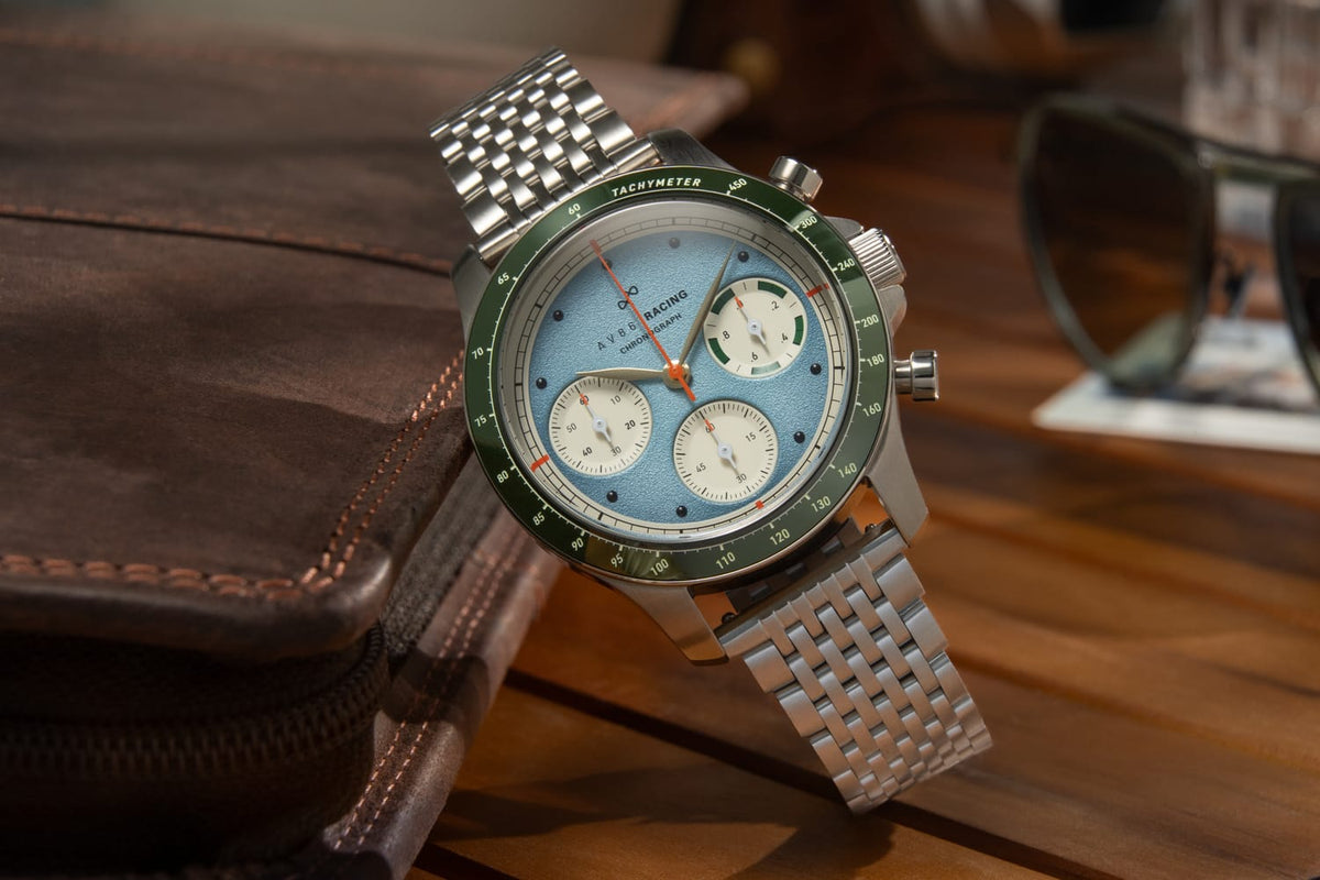 AV86 1960 Racing Chronograph, Steel / Sky Blue & Green quartz chronograph with blue dial, green tachymeter bezel, three subdials, and steel bracelet beside a leather case