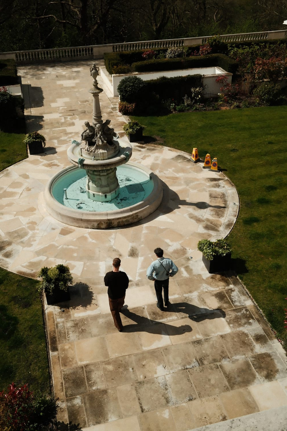 Two men walk along a sunlit stone pathway beside a decorative fountain, surrounded by manicured greenery and garden hedges, evoking a serene and refined outdoor ambiance ideal for appreciating timeless Scandinavian design such as the 1564 Chronodate Automatic, Steel / Blue by AV86.
