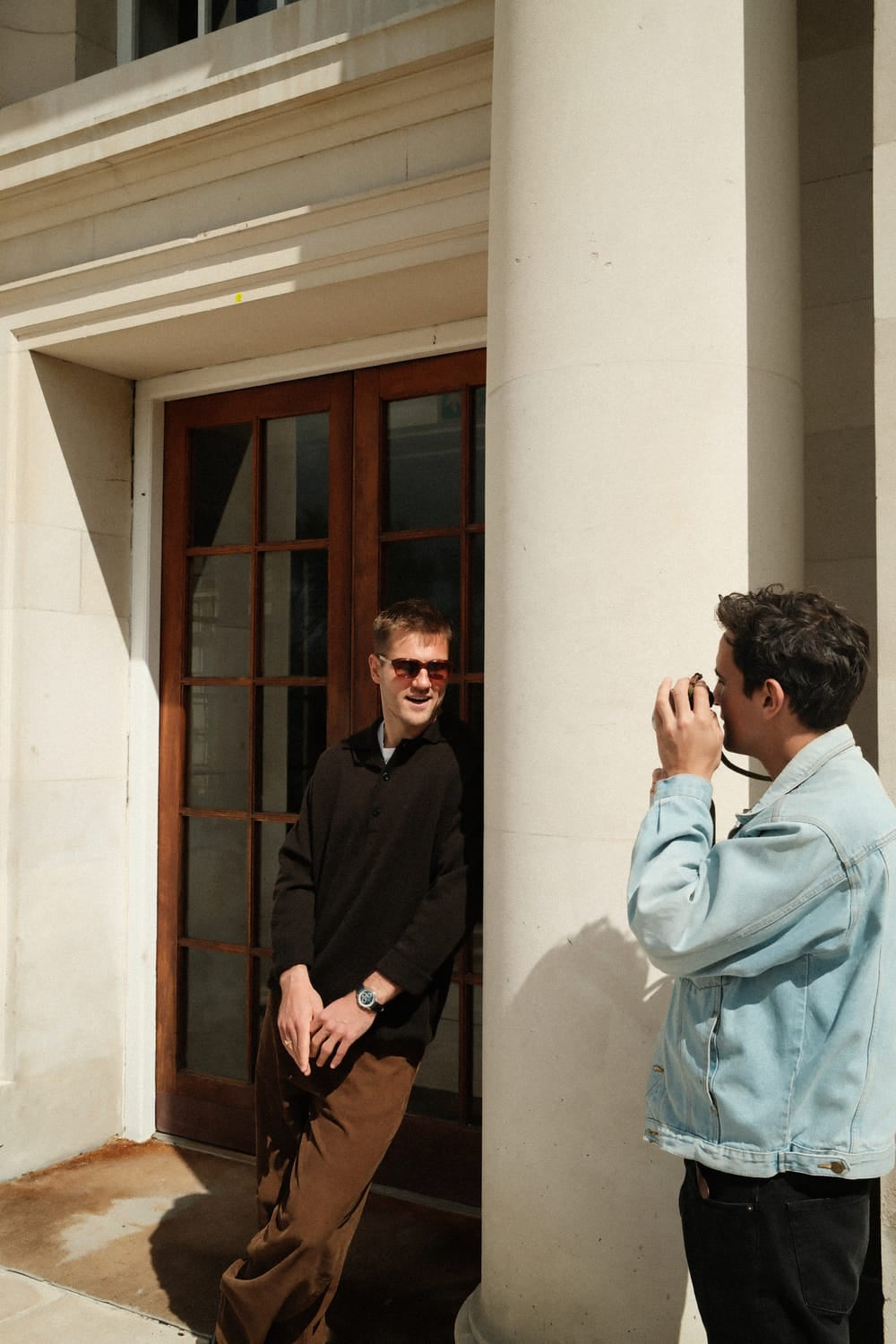 Two men engage in a casual photoshoot outside a classical building with stone columns; one wears a black sweater and brown trousers showcasing the 1564 Chronodate Automatic by AV86, embodying refined Nordic minimalism and vintage-inspired chronograph design.