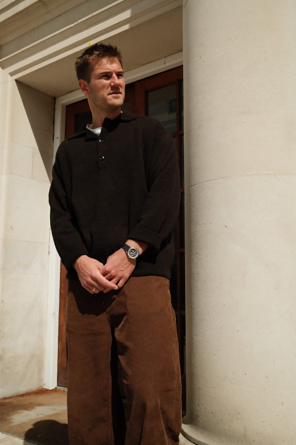 A man stands in front of a classical stone building wearing a dark knit sweater and brown trousers, showcasing the 1564 Chronodate Automatic by AV86 with its vintage-inspired chronograph dial and refined leather strap, embodying Danish design and Nordic minimalism in natural sunlight.