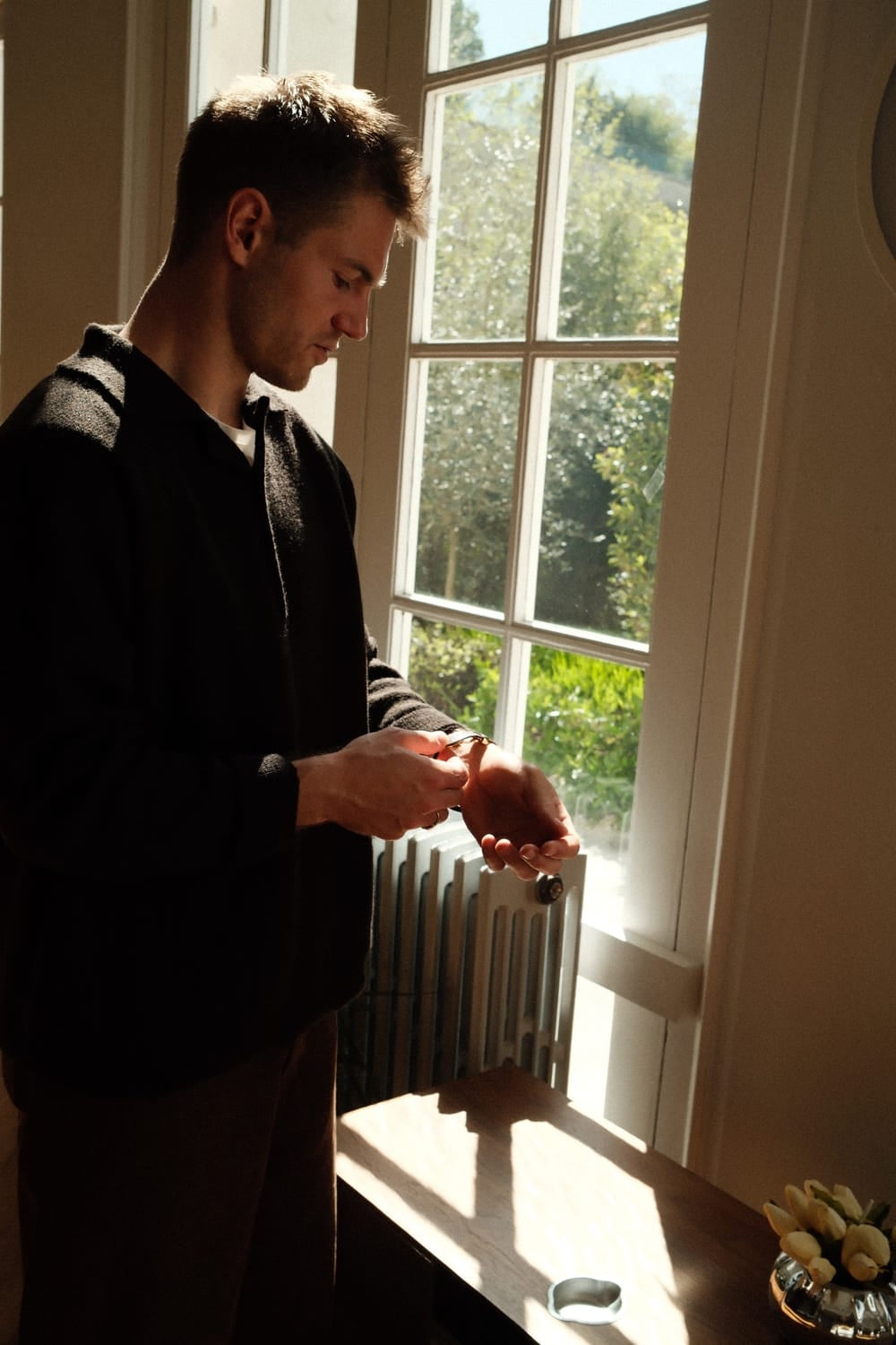 A man stands by a sunlit window adjusting a 1564 Chronodate Automatic, Steel / White on his wrist, dressed in a dark sweater, evoking a calm, refined Nordic ambiance with natural light highlighting the minimalist interior.