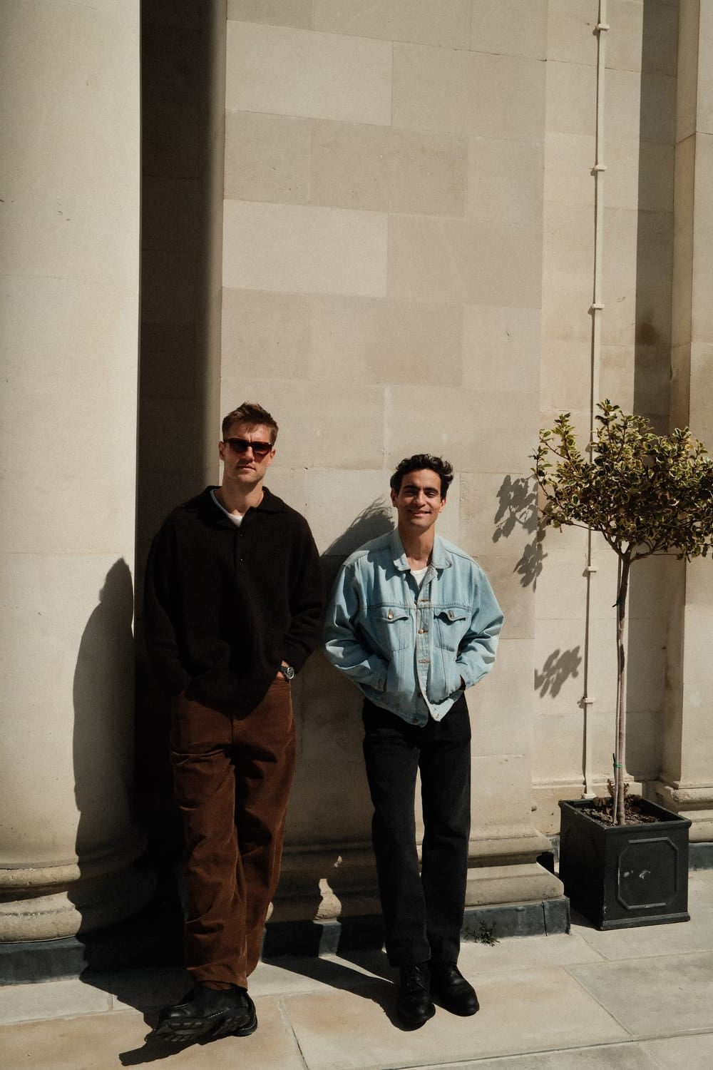 Two men stand against a stone column wall bathed in sunlight, one wearing relaxed brown trousers and a black sweater, the other in black pants and a light denim jacket, the first showcasing the 1564 Chronodate Automatic by AV86 with its vintage-inspired chronograph dial, polished stainless steel case, sapphire crystal, and leather strap, embodying refined Danish design and Nordic minimalism in a serene urban setting.