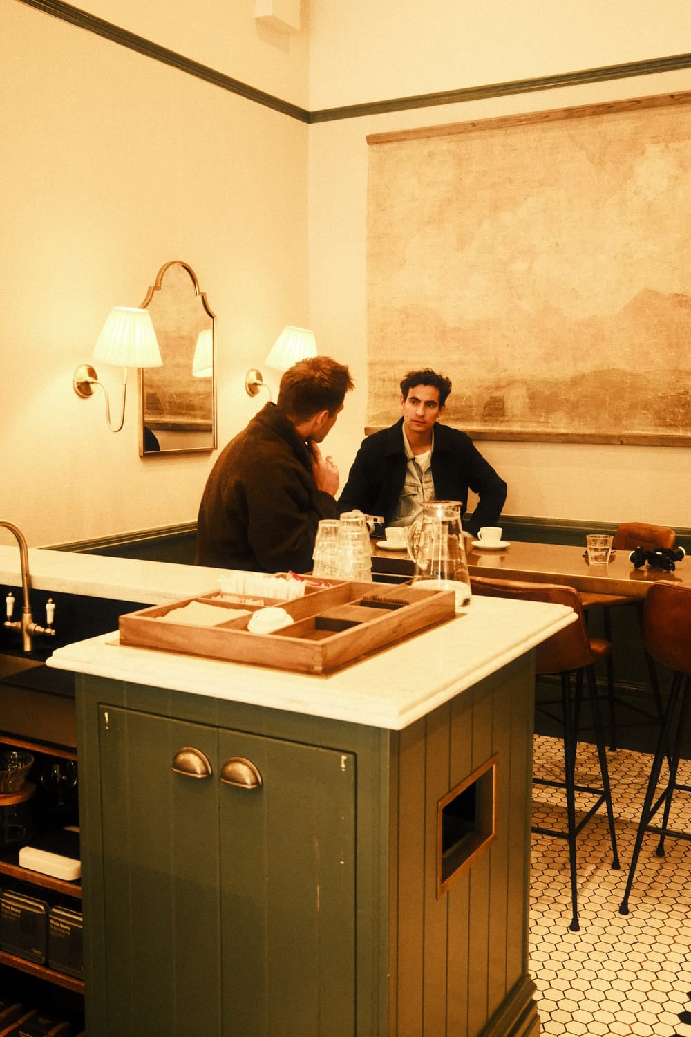 Two men engage in conversation in a warmly lit café with vintage decor, one wearing the 1564 Chronodate Automatic by AV86, a luxury chronograph with sapphire crystal and leather strap, reflecting refined Scandinavian craftsmanship and Nordic minimalism.