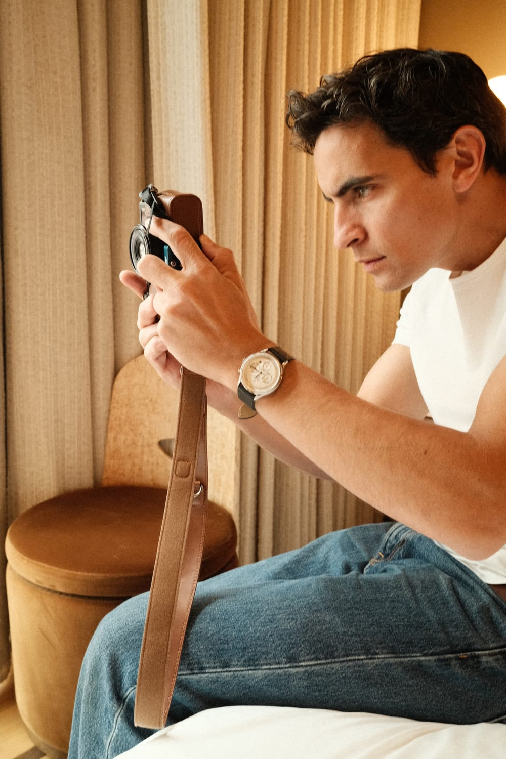 A man dressed casually in a white t-shirt and jeans focuses intently while handling a vintage camera, wearing the 1564 Chronodate Automatic by AV86, a refined chronograph with a detailed dial and smooth black leather strap, evoking Scandinavian design and understated luxury.