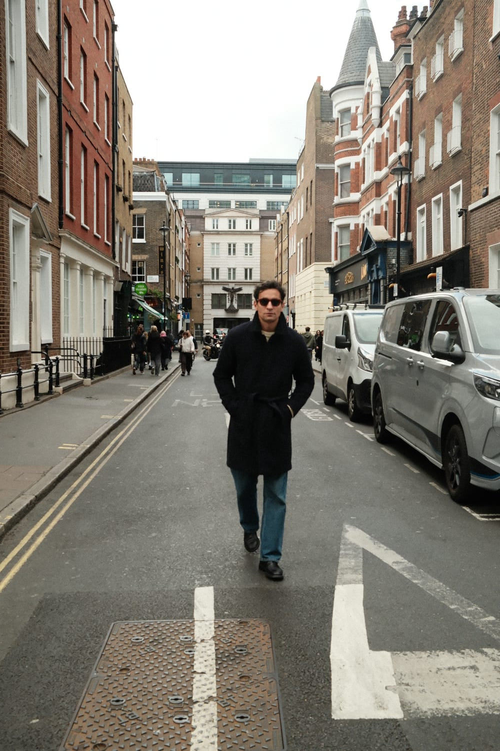 A man walks confidently down a city street wearing a dark overcoat and sunglasses, subtly showcasing the 1564 Chronodate Automatic by AV86, a vintage-inspired chronograph with a polished case, sapphire crystal, and black leather strap, embodying refined Danish design and Nordic minimalism.