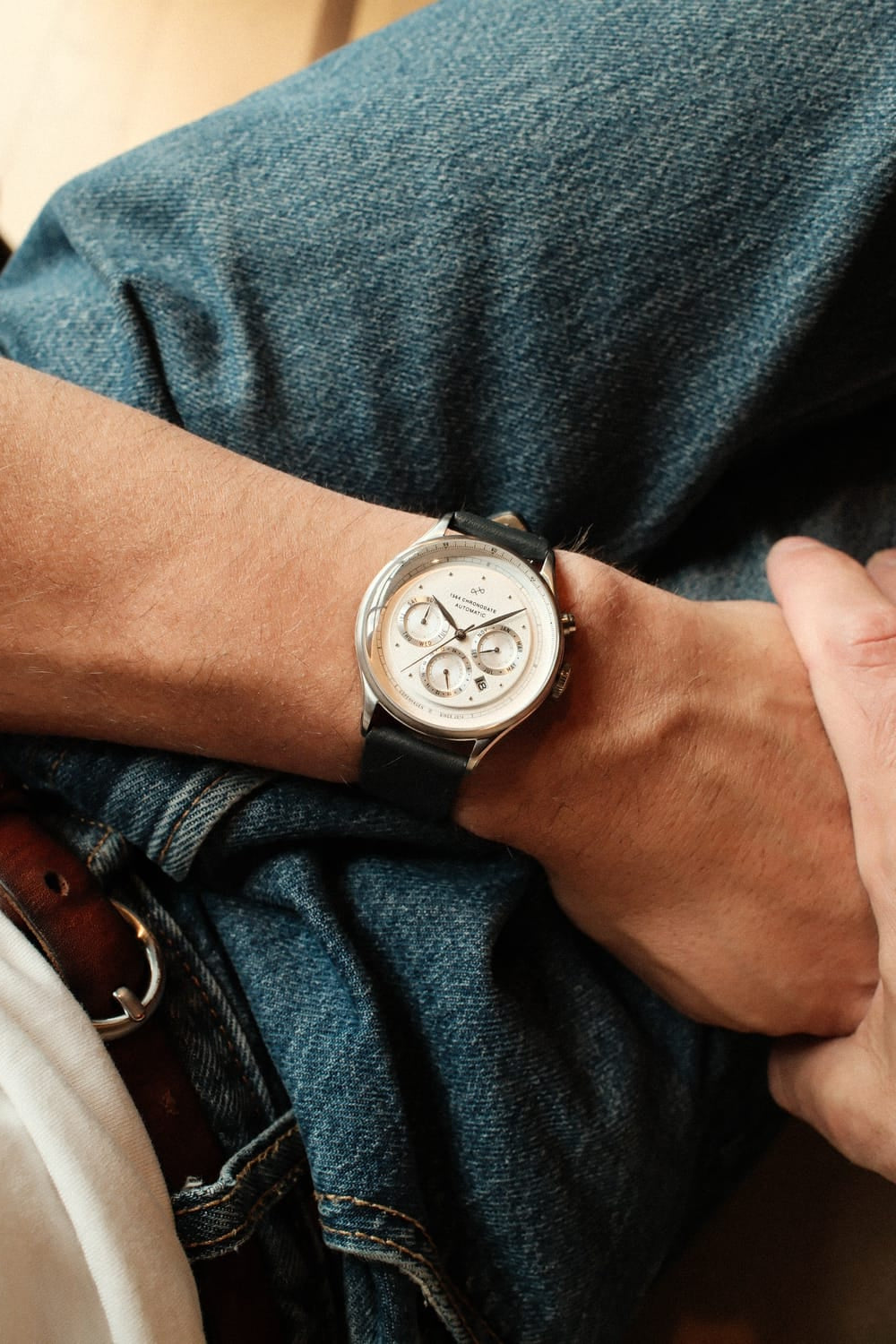 1564 Chronodate Automatic by AV86 features a vintage-inspired chronograph with a white dial, triple subdials, polished case, sapphire crystal, and black leather strap, worn casually with denim for a relaxed yet refined look embodying Danish design and Nordic minimalism.