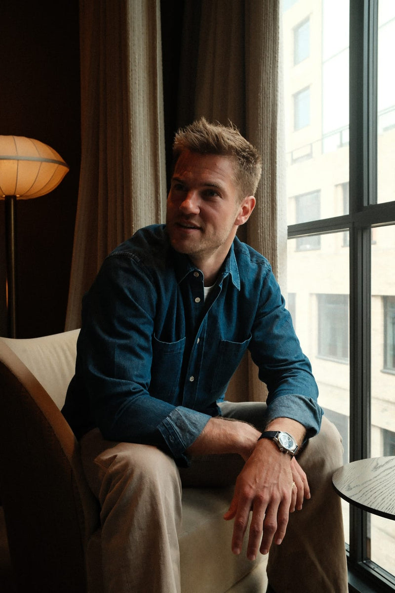 A man wearing a 1564 Chronodate Automatic, Steel / Blue by AV86 seated indoors by a window, dressed in a denim shirt and beige trousers, exuding relaxed Nordic minimalism and understated elegance.