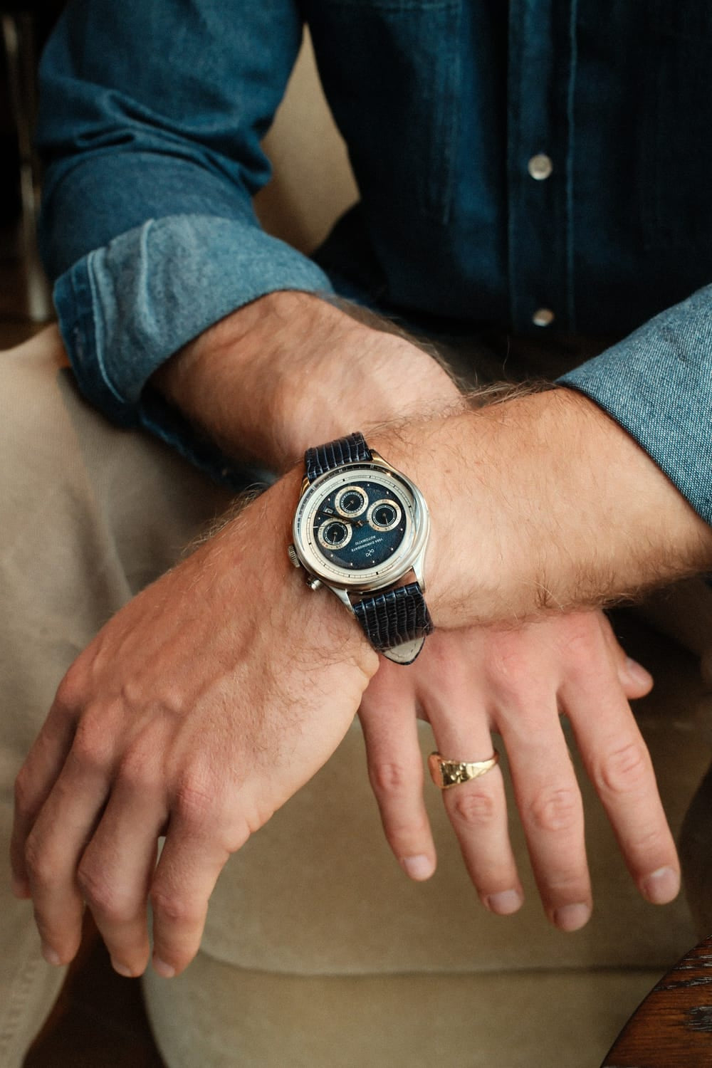 A man dressed in a rolled-up denim shirt and beige trousers casually leans forward, wearing the 1564 Chronodate Automatic, Steel / Blue by AV86 with its polished case and textured blue leather strap, embodying refined Scandinavian style and understated luxury.