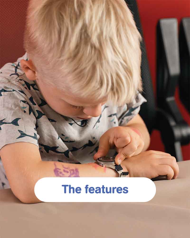 Blond child examining an AV86 wristwatch on an adult wrist at a table, with a rounded The features button overlaid against a red backdrop