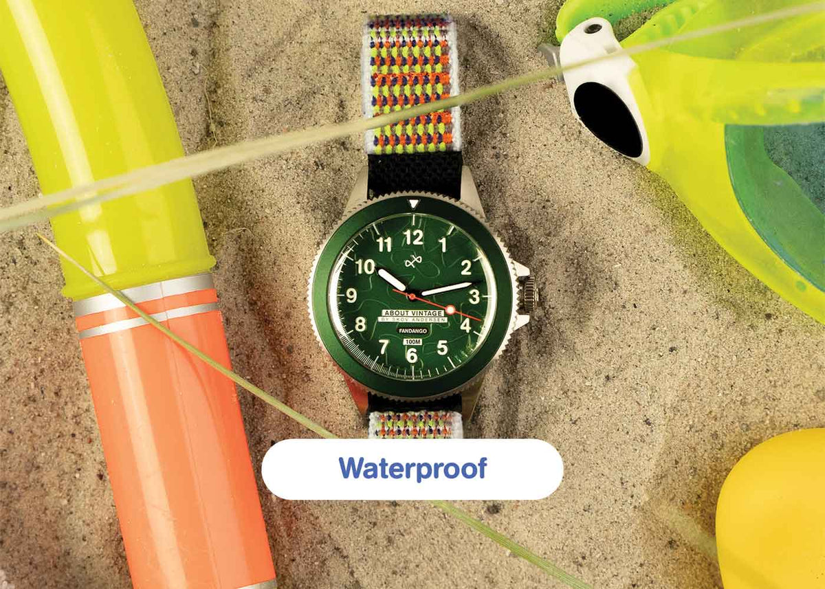AV86 diver wristwatch with green Arabic numeral dial, black bezel and patterned woven strap on sand beside colorful pool toys, labeled Waterproof