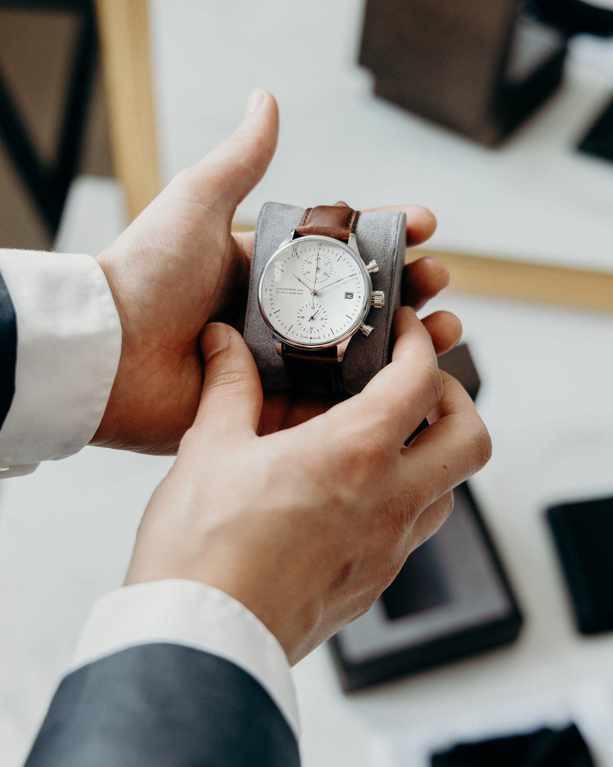 Chronograph by AV86 with sapphire crystal, minimalist dial, date function, and brown leather strap held in hands over a tray