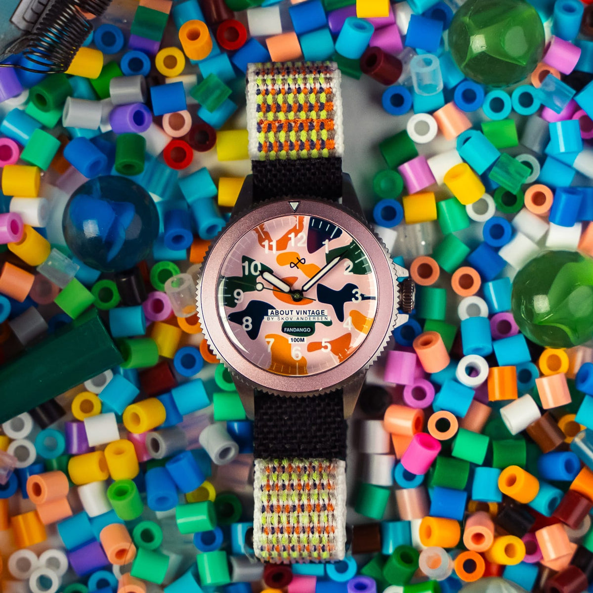 AV86 automatic watch with warm-toned dial, bold Arabic numerals, dark case and woven multicolor strap surrounded by colorful beads