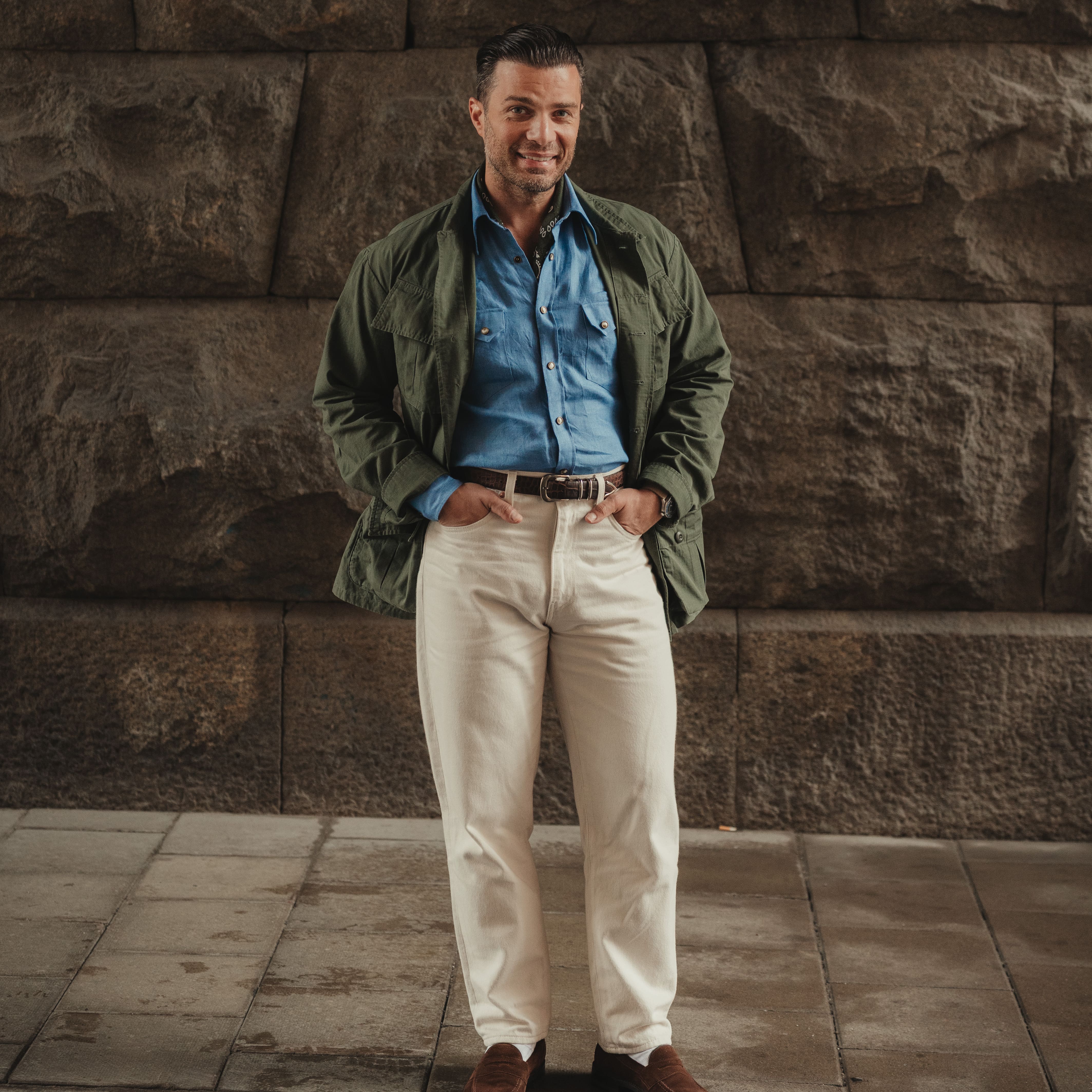 Man in casual outdoor attire with green jacket, blue shirt, beige trousers, and brown shoes against textured stone wall background
