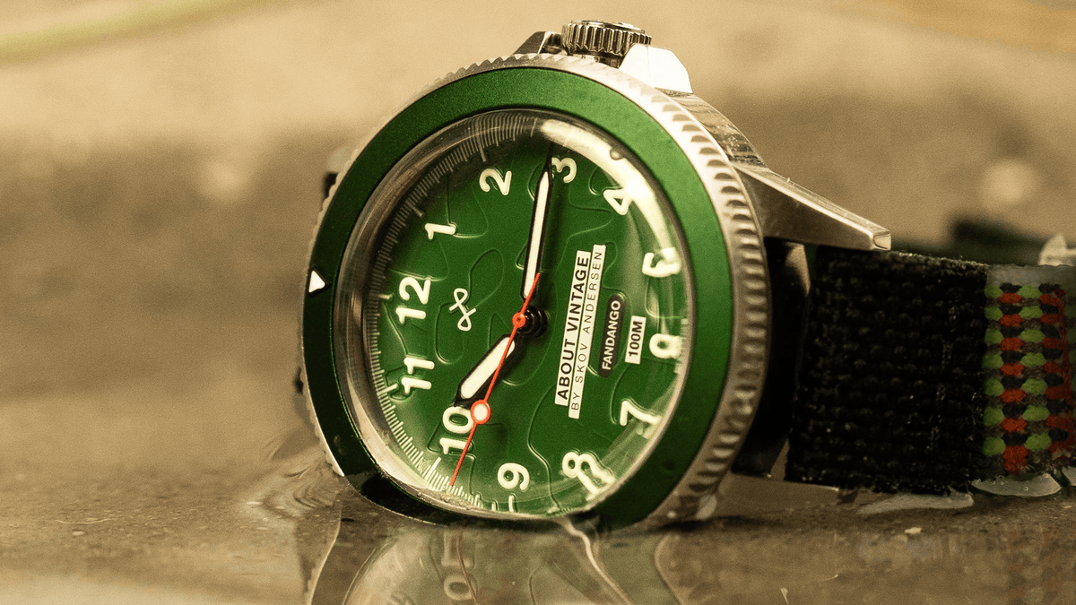 AV86 analog wristwatch with green dial, luminous Arabic numerals, polished steel case, black bezel, and black woven strap with colored accents on a reflective surface