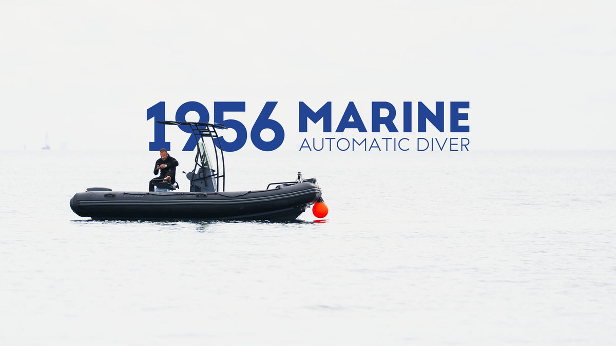 Small rigid inflatable boat on calm pale water beneath bold blue lettering reading 1956 Marine Automatic Diver.