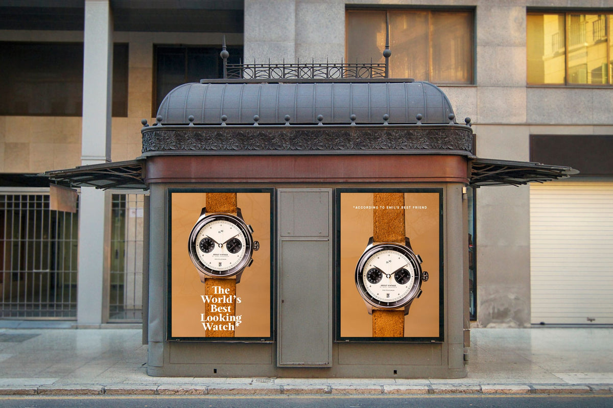 Street advertising kiosk displaying two matching chronograph watches with cream dials and brown leather straps against an urban building facade