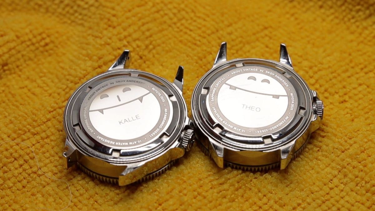 Two AV86 wristwatches with polished silver cases and white casebacks labeled Julie and Theo, resting on textured mustard fabric
