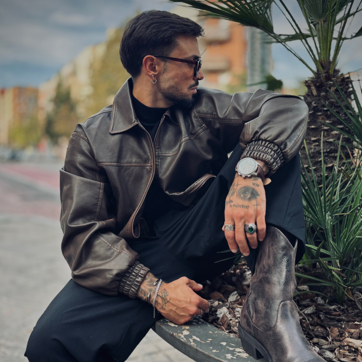 Contemporary urban style featuring a man in leather jacket, sunglasses, and boots with a wristwatch in a relaxed outdoor setting