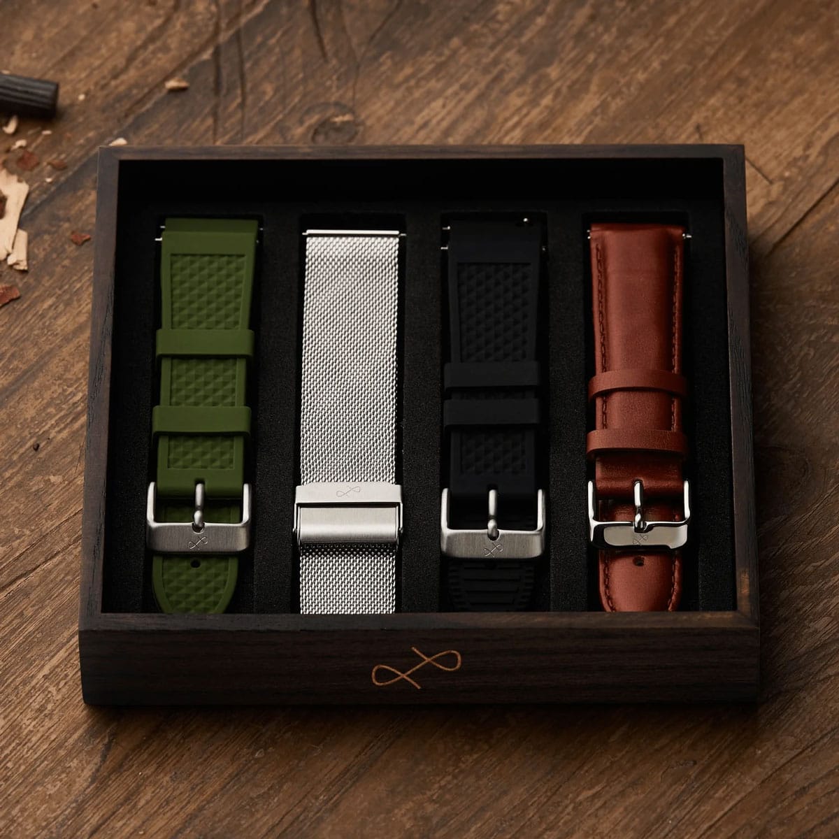 Four interchangeable watch straps in green, mesh metal, black and brown leather arranged in a wooden presentation box on a rustic table