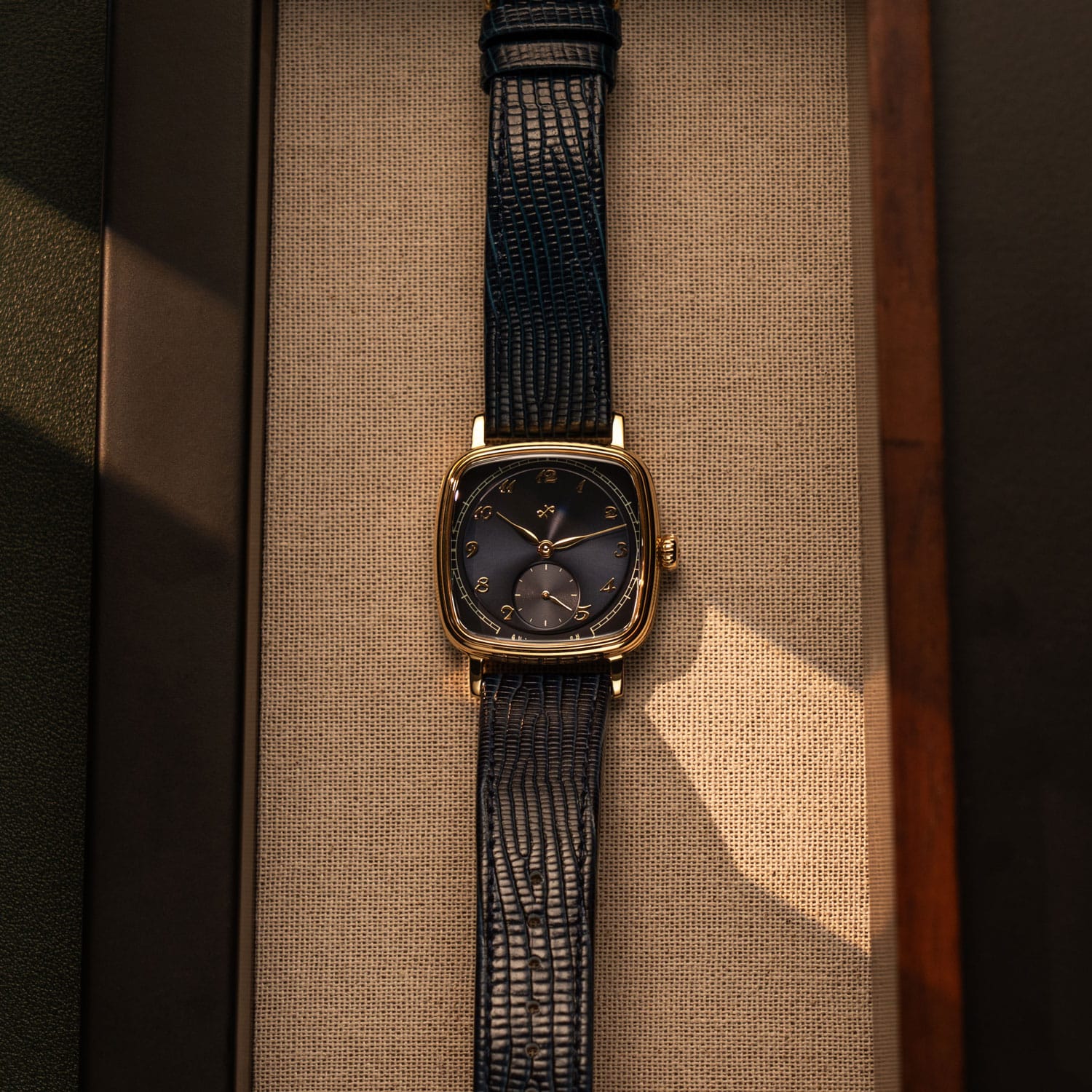 Vintage-inspired wristwatch with square gold case, dark dial, gold numerals, and textured black leather strap on beige fabric.