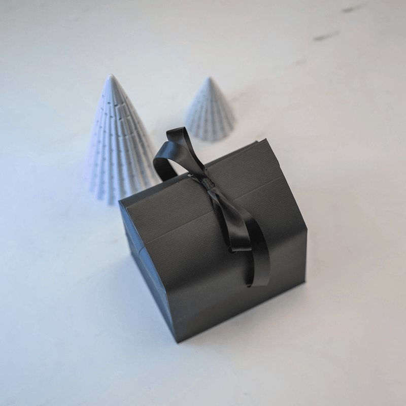 Gaveindpakning fra AV86 med en elegant sort æske bundet med et satinbånd ved siden af to minimalistiske papirkegler på en hvid baggrund.