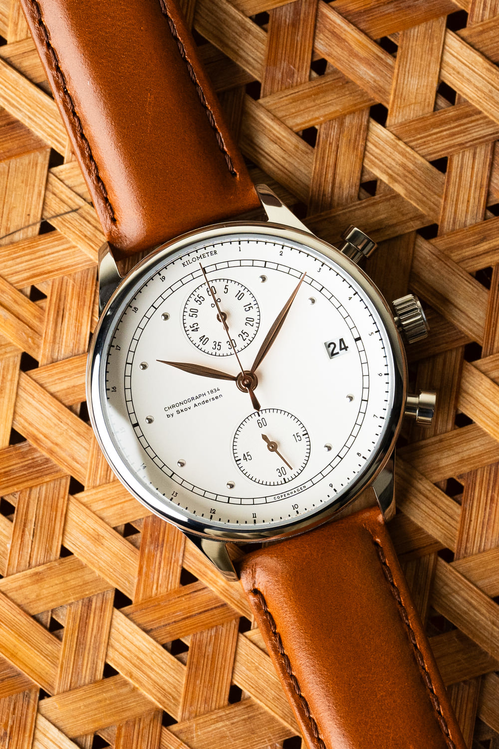 1834 Chronograph by AV86 luxury chronograph with sapphire crystal, white dial, steel case, and brown leather strap on wicker background