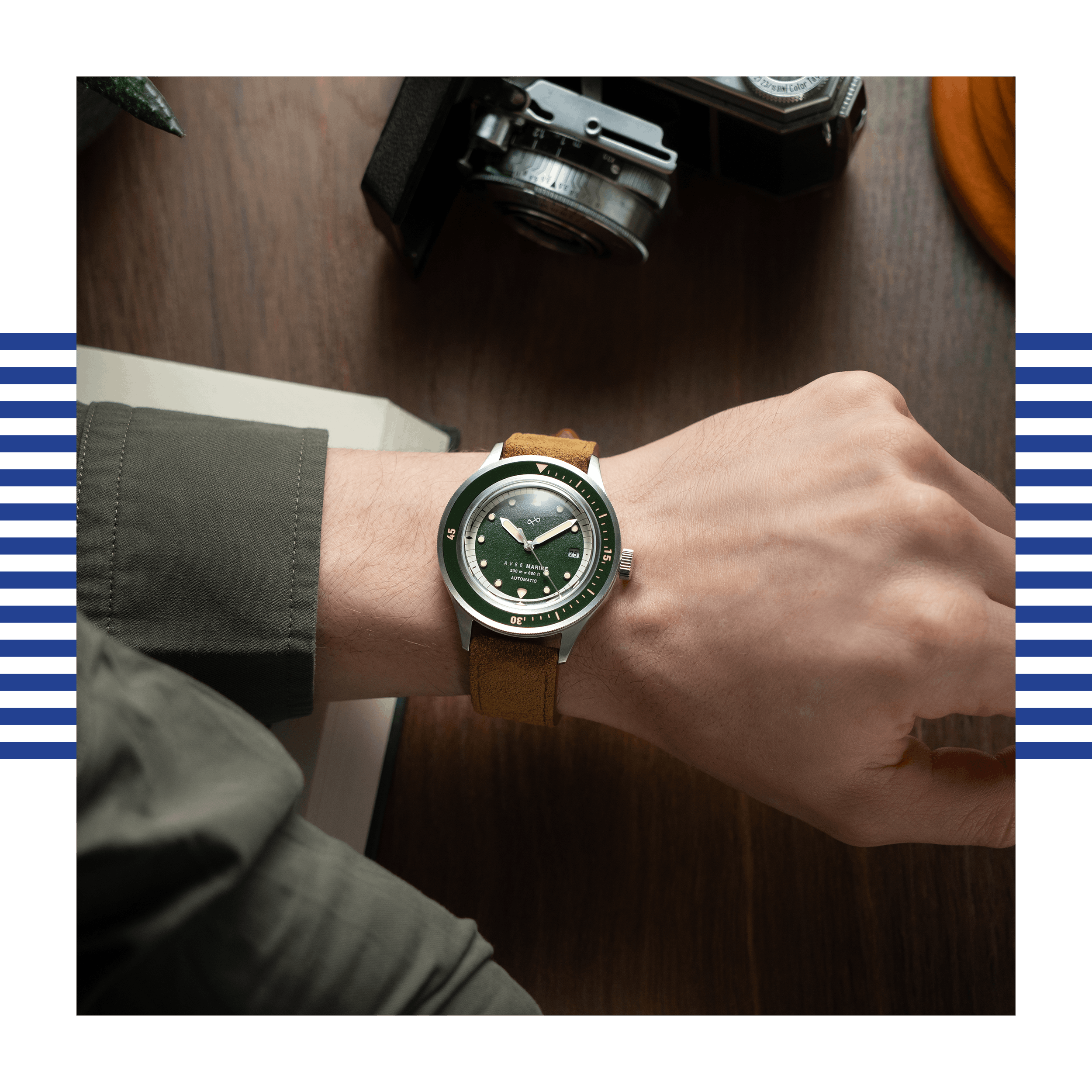 Wrist wearing a stainless steel watch with a deep green dial and brown leather strap on a wooden desk background.