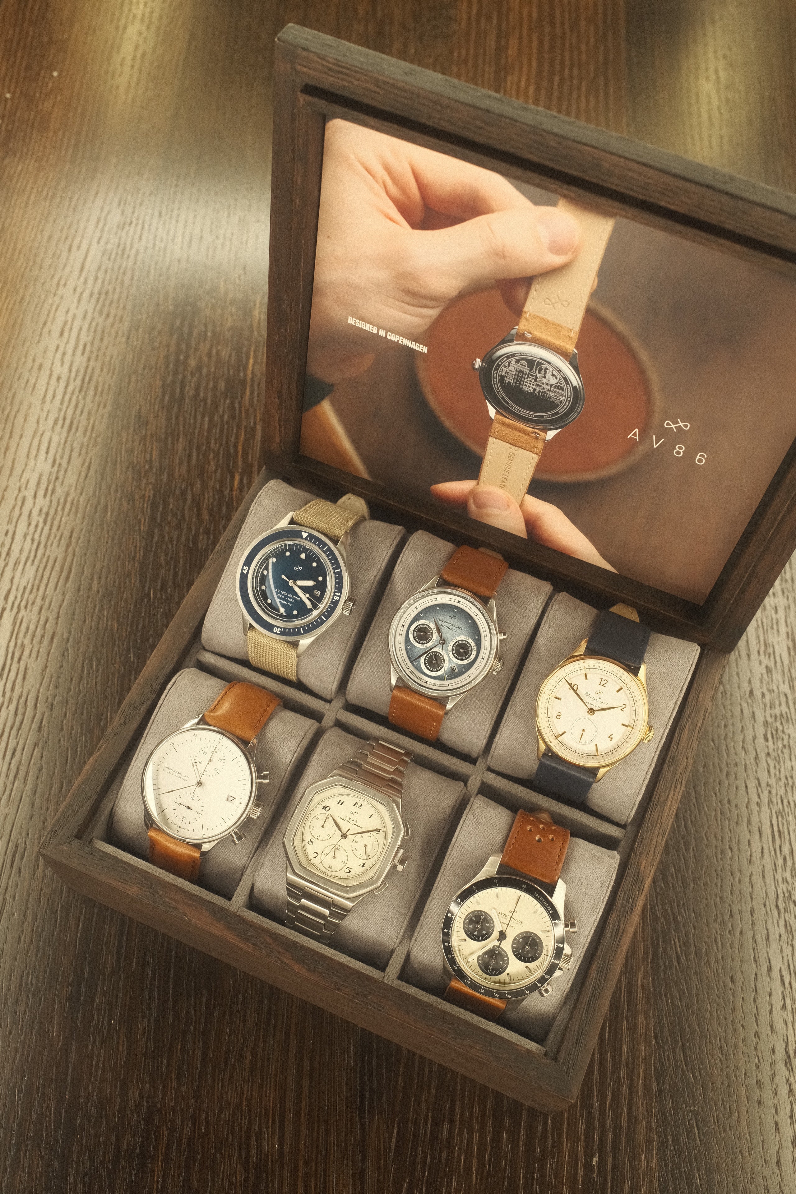 Wooden display box with six diverse wristwatches featuring leather, fabric, and metal straps arranged on soft cushions.