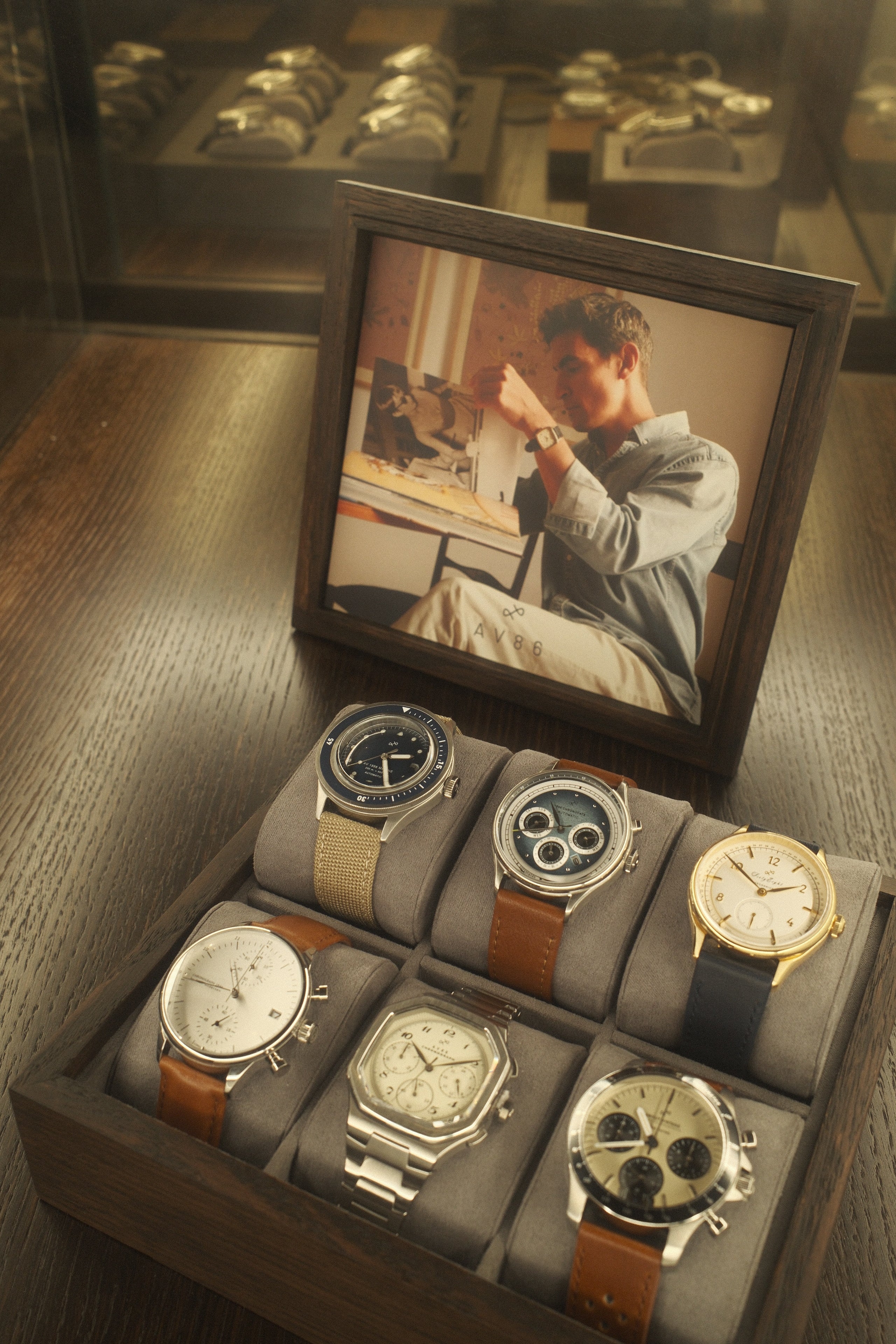 Wooden display box with six wristwatches featuring various strap materials and dial designs beside a framed portrait of a man wearing a watch.