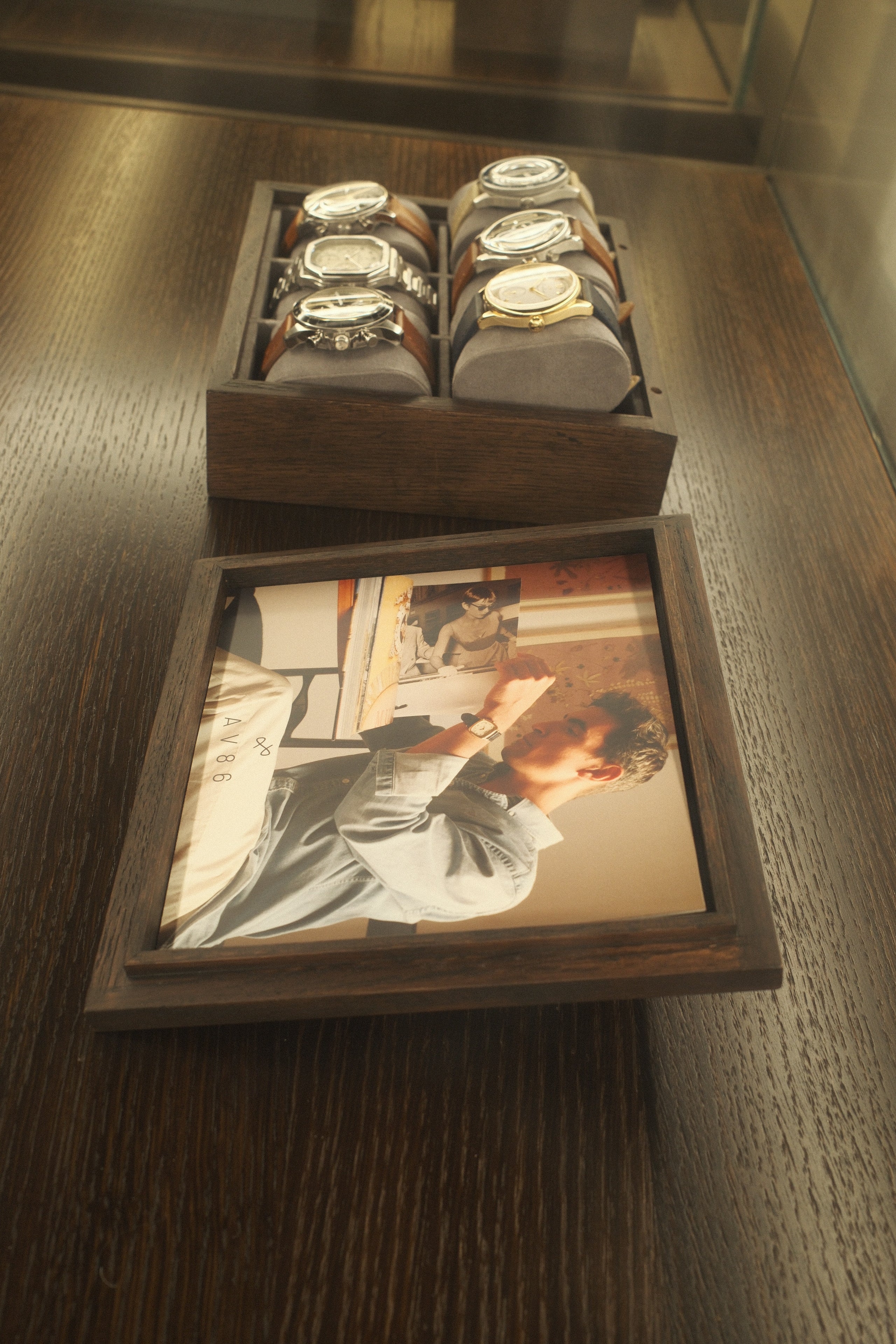 Wooden box displaying six wristwatches with metal and leather straps on soft cushions beside a framed photo of a man wearing a watch
