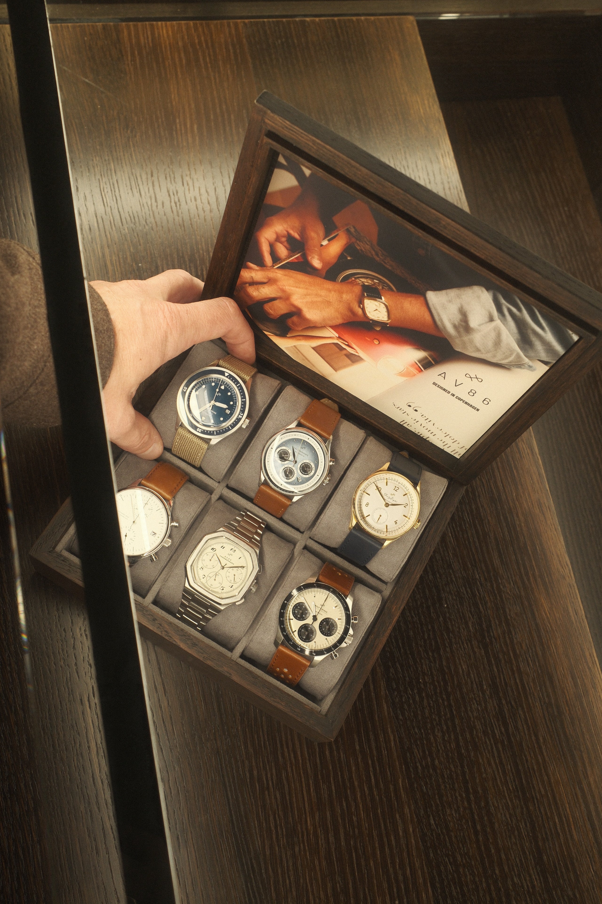 Wooden box holding six wristwatches with diverse designs including metal, leather, and fabric straps on a dark wood surface.