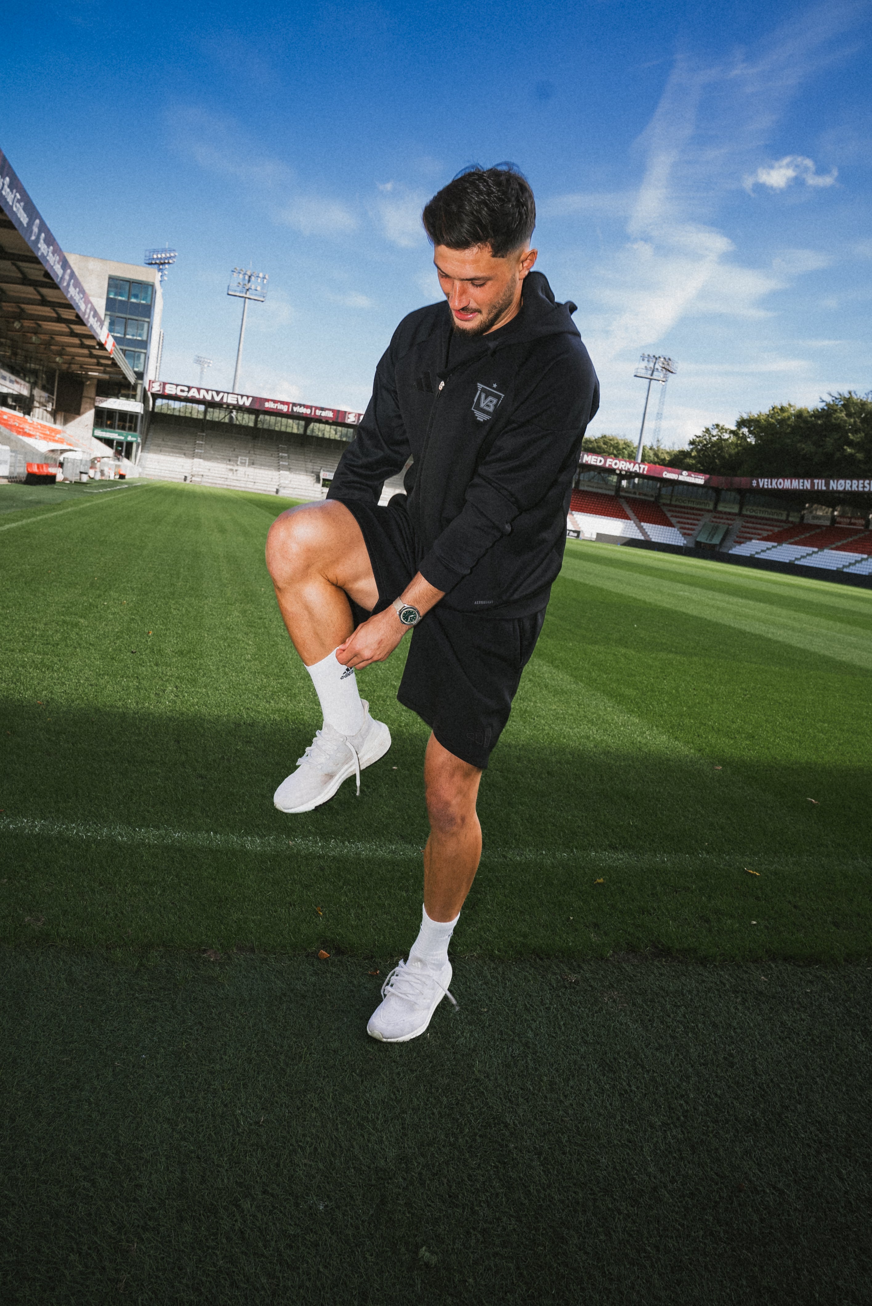 Athletic man in black hoodie and shorts adjusting his shoe on a green sports field under clear blue sky with casual sporty vibe