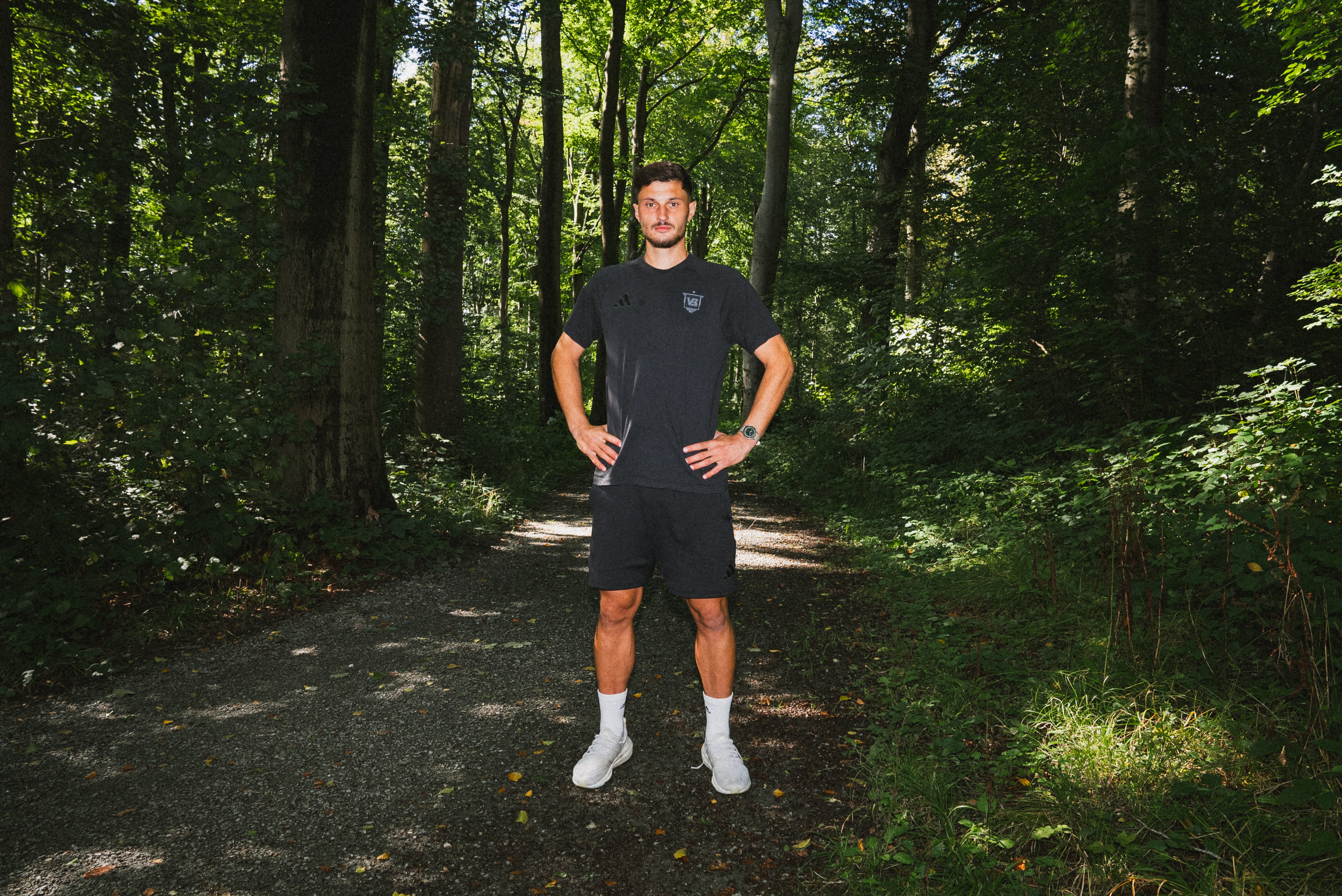 Young man standing confidently on forest trail wearing casual athletic attire and AV86 wristwatch, natural outdoor setting