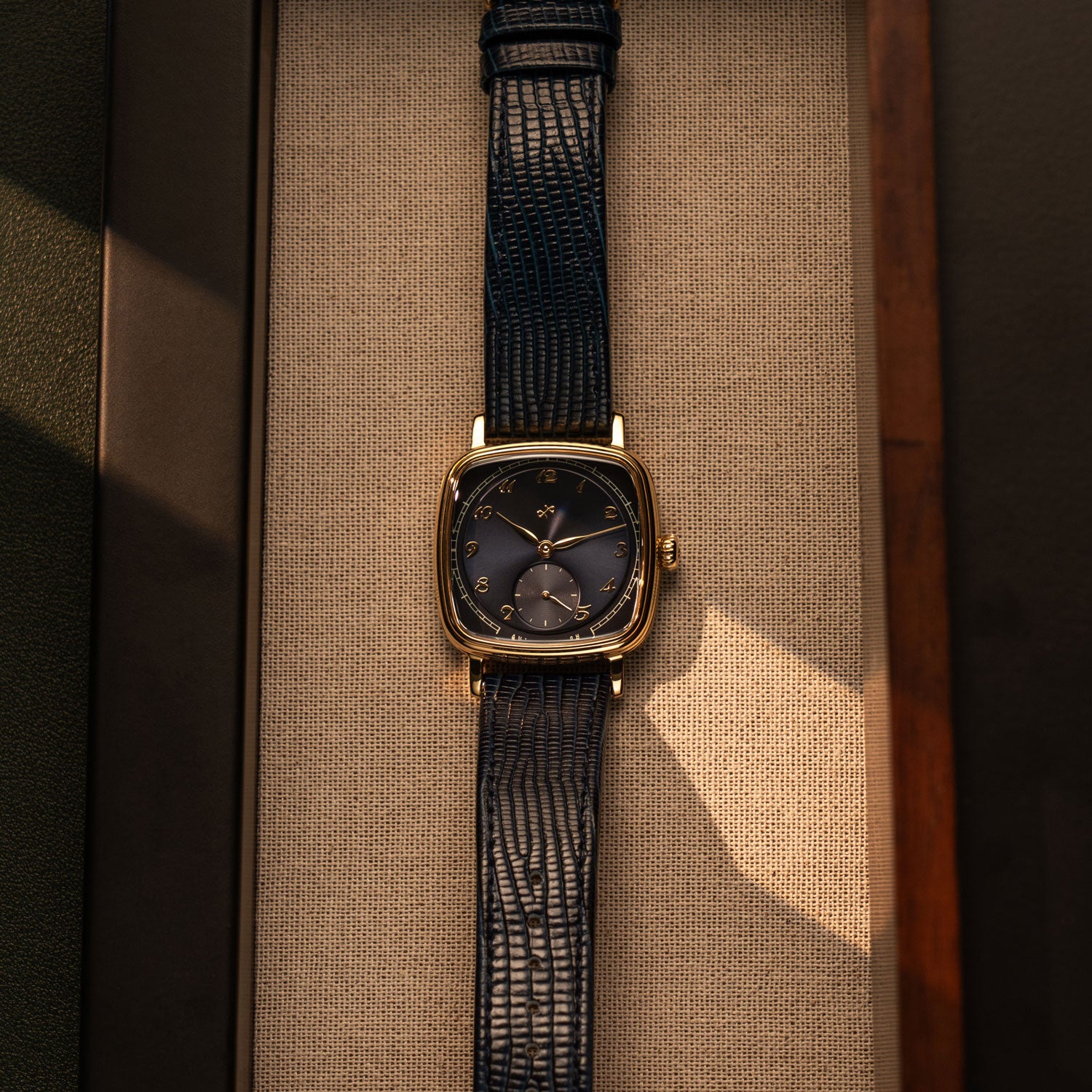 Vintage-inspired wristwatch with square gold case, dark dial, gold numerals, and textured black leather strap on beige fabric.