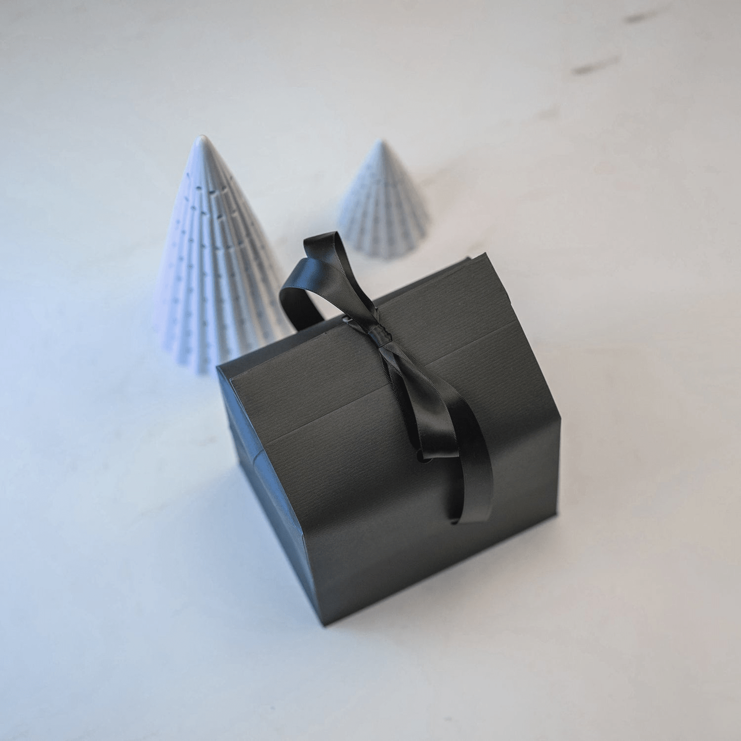 Gift wrapping by AV86 featuring a sleek black box tied with a satin ribbon beside two minimalist paper cones on white surface.